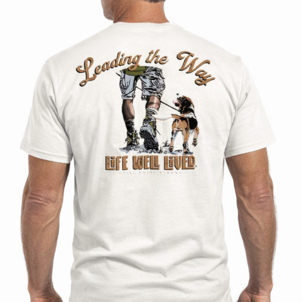 Back view of white Leading the Way T‑shirt featuring illustration of person walking a beagle with Leading the Way text above and Life Well Lived Still Going Strong branding below.