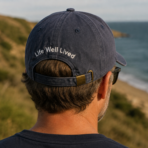 A man viewed from behind wearing a navy blue baseball cap with 'Life Well Lived' embroidered in white on the back, standing on a grassy cliff overlooking the ocean.