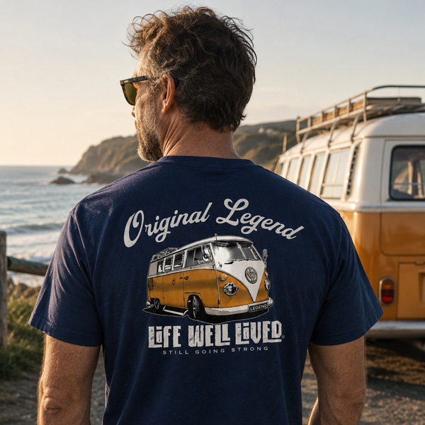 A man standing on a beach facing away from the camera, wearing a navy blue t-shirt with a large graphic print of a vintage orange and white van and the text 'Original Legend' and 'Life Well Lived'. A classic orange and white van is parked in the background near the ocean.