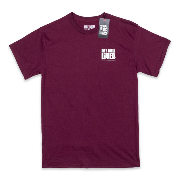 Mens Graphic Tee shirt, in a maroon colour, it is a back print T with a small chest print logo, the design on the Tee is of a classic route 66 sign shape, with the words Life Well Lived in a block font inside the sign.