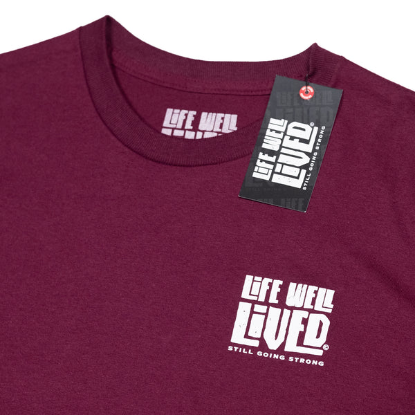 Mens Graphic Tee shirt, in a maroon colour, it is a back print T with a small chest print logo, the design on the Tee is of a classic route 66 sign shape, with the words Life Well Lived in a block font inside the sign.