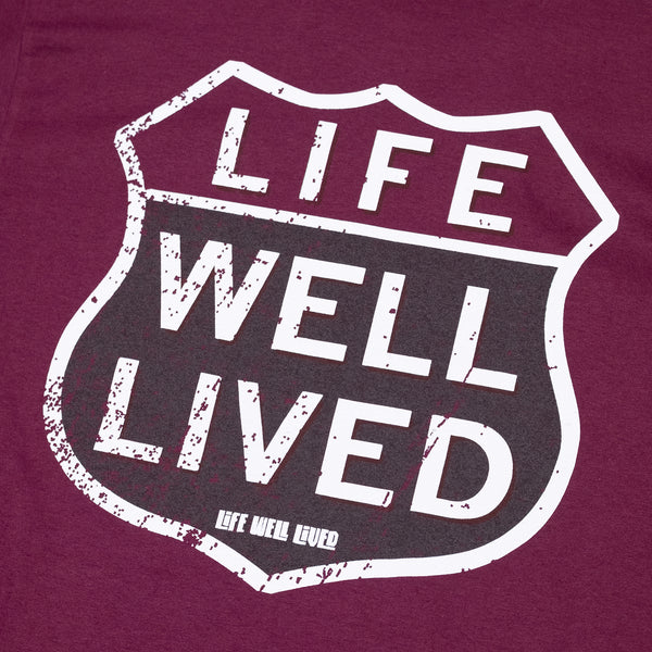 Mens Graphic Tee shirt, in a maroon colour, it is a back print T with a small chest print logo, the design on the Tee is of a classic route 66 sign shape, with the words Life Well Lived in a block font inside the sign.