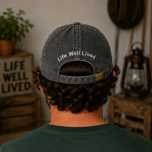 A person viewed from behind wearing a dark washed baseball cap with the words 'Life Well Lived' embroidered on the back, in a rustic indoor setting with wooden crates and a lantern in the background.