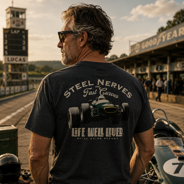 A man stands on a racetrack wearing a dark charcoal gray t-shirt with a vintage green and white race car graphic on the back, featuring the text 'STEEL NERVES Fast Gears' and 'LIFE WELL LIVED'.