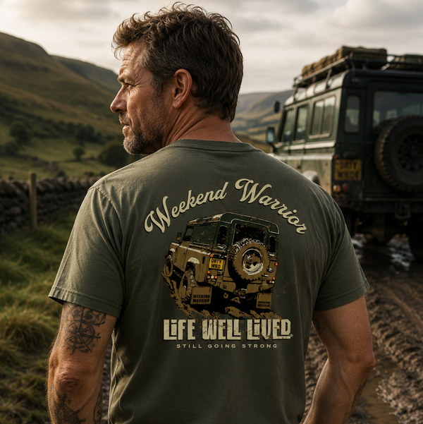 A man stands outdoors on a muddy road wearing an olive green graphic t-shirt, viewed from behind. The shirt features a large graphic of a military-style jeep with the text 'Weekend Warrior' and 'Life Will Lived.