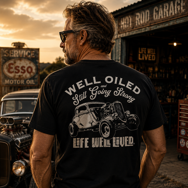 A man viewed from behind wearing a black graphic t-shirt with a white hot rod car design and the text 'WELL OILED Still Going Strong LIFE WE'LL LIVED'. The background depicts a warm sunset at a vintage auto repair shop with an Esso sign and a classic car.