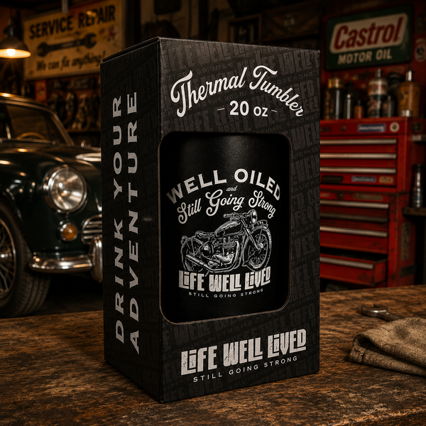 A black 20 oz Thermal Tumbler mug with a white graphic of a motorcycle and the text 'WELL OILED and Still Going Strong' displayed inside its branded black packaging box on a wooden workbench with a garage background.