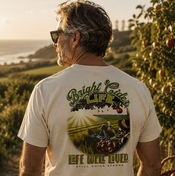 A man wearing a cream-colored t-shirt with a large green and brown graphic on the back, standing in an outdoor vineyard at sunset.