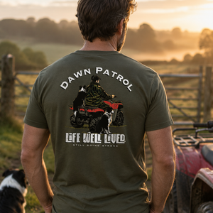 A man viewed from behind wearing an olive green t-shirt with a large graphic on the back that reads 'DAWN PATROL' and 'LIFE WELL LIVED' with an ATV and dog illustration. He is standing outdoors in a rural setting at sunset next to an ATV, with a dog sitting nearby.