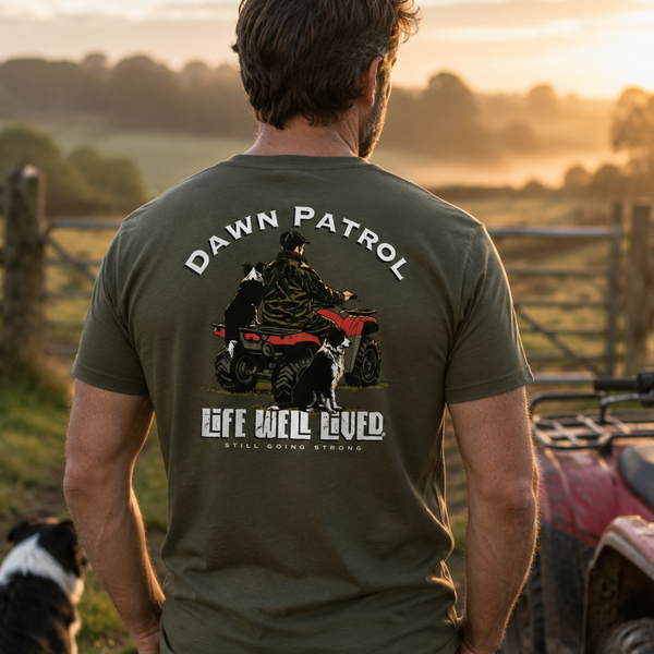 A man viewed from behind wearing an olive green t-shirt with a large graphic on the back that reads 'DAWN PATROL' and 'LIFE WELL LIVED' with an ATV and dog illustration. He is standing outdoors in a rural setting at sunset next to an ATV, with a dog sitting nearby.
