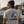 A man wearing a white graphic t-shirt with a blue and black tuna fish design and the text 'DEEP BLUE GURU' and 'LIFE WELL LIVED' on the back, standing on a dock with boats and water in the background.