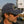 A man in profile wearing a faded navy blue baseball cap with a 'Steel Motors' patch and dark sunglasses, photographed outdoors at a race track with a green race car and a 'GIRLING' sign in the blurred background.