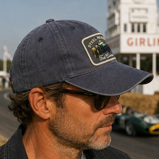 A man in profile wearing a faded navy blue baseball cap with a 'Steel Motors' patch and dark sunglasses, photographed outdoors at a race track with a green race car and a 'GIRLING' sign in the blurred background.