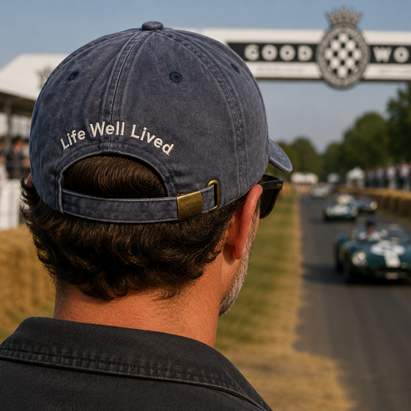 A man viewed from behind wearing a navy blue baseball cap with 'Life Well Lived' embroidered in white on the back, standing at Goodwood Racecourse with vintage race cars and the Goodwood Arch in the background.