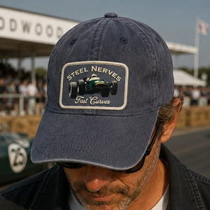 A man wearing a navy blue baseball cap with an embroidered patch that reads 'STEEL NERVES Fast Gears' and features a green and black race car graphic. The background shows a blurred racetrack scene with a vintage green race car and a crowd under a 'GOODWOOD' sign.
