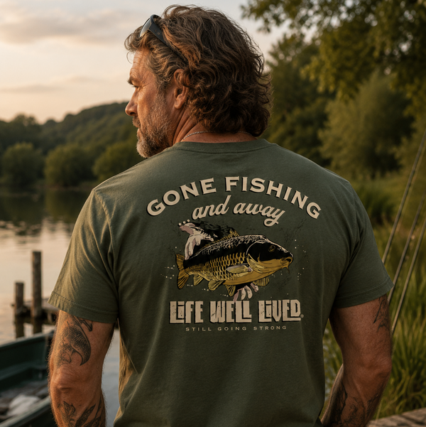A man standing outdoors by a lake at sunset, shown from the back wearing an olive green t-shirt with a large graphic print that reads 'GONE FISHING and away' and 'LIFE WILL BEVERED. WILLLI BURNED OVER'.