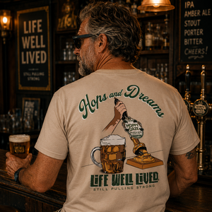 A man sitting at a bar, viewed from behind, wearing a beige graphic t-shirt with a large beer-themed print on the back. The design features the text 'Hops and Dreams' and 'Life Well Lived' in green and black, alongside illustrations of a beer mug, hops, and a pint glass.