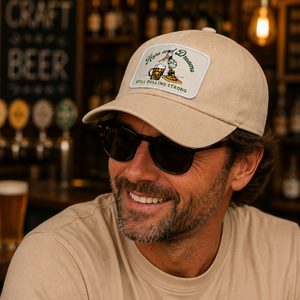 A smiling man wearing a beige baseball cap and matching beige t-shirt in a warm, dimly lit bar setting. The cap features a white patch with a green and gold design that reads 'Sycamore Brewing Co.