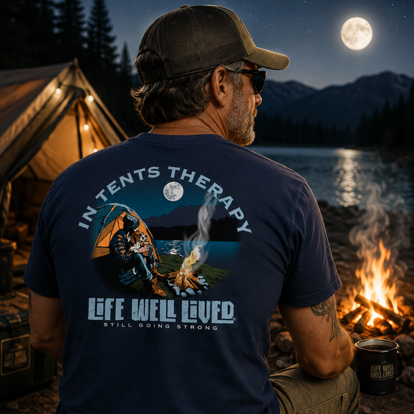 A man sits with his back to the camera at a lakeside campsite at night, wearing a navy blue graphic t-shirt. The shirt features a large back print with a tent, campfire, moon, and the text 'IN TENTS THERAPY' and 'LIFE WELL LIVED'.
