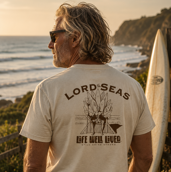 A man standing on a beach path at sunset, viewed from behind, wearing a cream-colored t-shirt with a dark brown graphic that reads 'LORD SEAS' and 'LIFE WELL LOVED'. A surfboard leans against a cliff in the background.