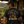 A man wearing a black graphic t-shirt with a large 'MEAT, FIRE, GLORY' and 'LIFE WILL BE LOVED' design on the back, standing in a rustic outdoor campsite setting with a grill and mountains in the background.