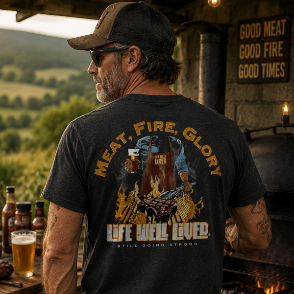 A man wearing a black graphic t-shirt with a large 'MEAT, FIRE, GLORY' and 'LIFE WILL BE LOVED' design on the back, standing in a rustic outdoor campsite setting with a grill and mountains in the background.