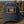 A navy blue baseball cap with a rectangular embroidered patch on the front, placed on a wooden table with a blurred outdoor background. The patch reads 'Original Legend' and 'Still Going Strong' with a yellow and white van illustration.