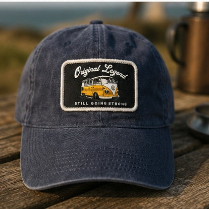 A navy blue baseball cap with a rectangular embroidered patch on the front, placed on a wooden table with a blurred outdoor background. The patch reads 'Original Legend' and 'Still Going Strong' with a yellow and white van illustration.