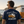 A man standing on a beach facing away from the camera, wearing a navy blue t-shirt with a large graphic print of a vintage orange and white van and the text 'Original Legend' and 'Life Well Lived'. A classic orange and white van is parked in the background near the ocean.