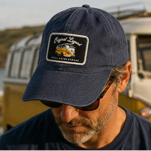 A man wearing a navy blue baseball cap with a rectangular embroidered patch that reads 'Original Legend' and 'Still Going Strong,' photographed outdoors in front of a vintage cream and yellow bus.