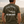 A man viewed from behind wearing an olive green t-shirt with a large back graphic that reads 'Peavy Climbers' and 'Life Well Lived. Wear Your Adventure'.