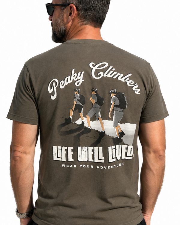 A man wearing a dark olive green t-shirt from the back, featuring a large graphic print of three climbers and the text 'Peaky Climbers' and 'LIFE WELL LIVED'.