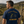 A man wearing a navy blue t-shirt with a yellow and black bicycle graphic and the text 'Professional Cyclopath' and 'Life Well Laid' on the back, standing outdoors next to a stone wall and a bicycle.
