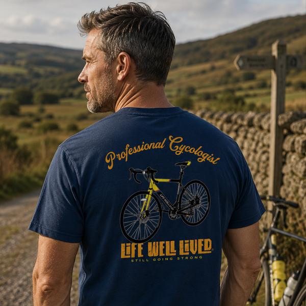 A man wearing a navy blue t-shirt with a yellow and black bicycle graphic and the text 'Professional Cyclopath' and 'Life Well Laid' on the back, standing outdoors next to a stone wall and a bicycle.
