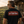 A man wearing a black t-shirt with a vintage-style red and white graphic on the back, standing outdoors in front of a rustic wooden building and a motorcycle at sunset.