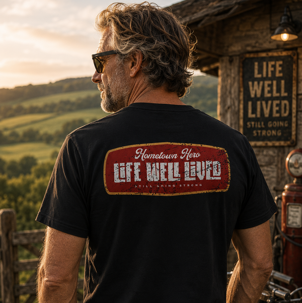 A man wearing a black t-shirt with a vintage-style red and white graphic on the back, standing outdoors in front of a rustic wooden building and a motorcycle at sunset.