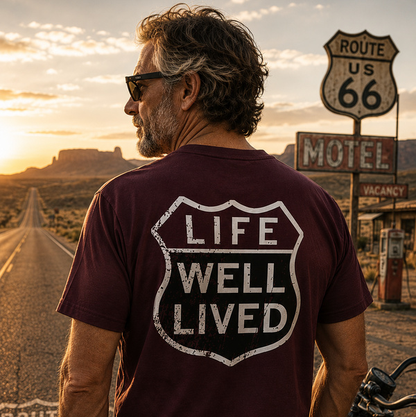 A man wearing a maroon t-shirt with a distressed white 'LIFE WELL LIVED' shield graphic on the back, standing on a desert road at sunset next to a motorcycle and Route 66 signs.