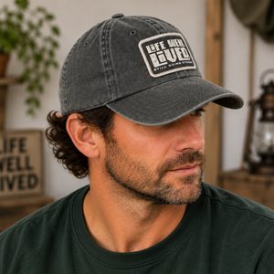 A man wearing a dark gray washed baseball cap with a white 'Life Well Lived' logo patch on the front, paired with a dark green t-shirt in a rustic indoor setting.