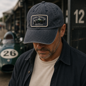 A man wearing a dark navy blue baseball cap with a 'Steel Nerves' patch, standing in front of a vintage race car and garage background.