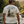 A man standing in a flowing river, viewed from behind, wearing a cream-colored t-shirt with a large back graphic that reads 'TIGHT LINES Calm Minds LIFE WELL LIVED WEAR YOUR ADVENTURE' with an illustration of a fisherman and a largemouth bass. The background shows a lush, green forest with warm golden-hour sunlight.