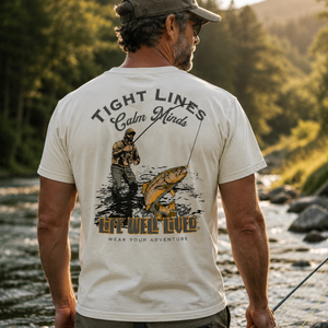 A man standing in a flowing river, viewed from behind, wearing a cream-colored t-shirt with a large back graphic that reads 'TIGHT LINES Calm Minds LIFE WELL LIVED WEAR YOUR ADVENTURE' with an illustration of a fisherman and a largemouth bass. The background shows a lush, green forest with warm golden-hour sunlight.