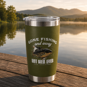 An olive green insulated tumbler with a clear lid sits on a wooden dock with a lake and mountains in the background. The tumbler has a graphic of a fishing rod and fish with the text 'GONE FISHING and away' and 'LIFE WILL LIVED.