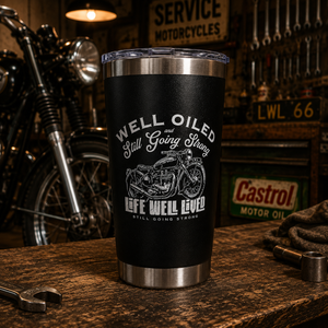 A black insulated tumbler with a clear lid sits on a wooden workbench in a motorcycle-themed garage setting. The tumbler has a brushed silver metal rim and base, and a white graphic print featuring a motorcycle and the text 'WELL OILED and Still Going Strong' and 'GEEW WELL LOVED'.