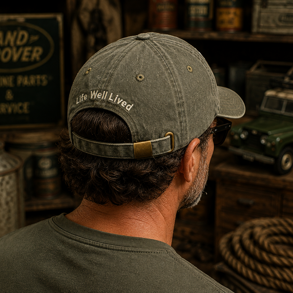 A man wearing a distressed olive green baseball cap with 'Life Well Lived' embroidered on the back, photographed from behind in a rustic workshop setting with Land Rover memorabilia.