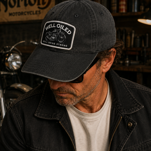 A man wearing a dark washed baseball cap with a white 'Well Oiled' patch, photographed in a motorcycle workshop with a Norton motorcycle in the background.