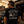 A man viewed from behind wearing a black graphic t-shirt with a white hot rod car design and the text 'WELL OILED Still Going Strong LIFE WE'LL LIVED'. The background depicts a warm sunset at a vintage auto repair shop with an Esso sign and a classic car.