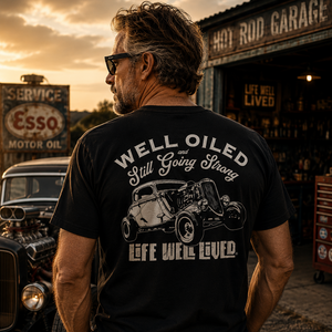 A man viewed from behind wearing a black graphic t-shirt with a white hot rod car design and the text 'WELL OILED Still Going Strong LIFE WE'LL LIVED'. The background depicts a warm sunset at a vintage auto repair shop with an Esso sign and a classic car.