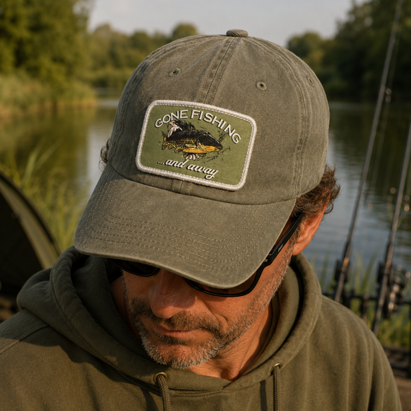 A man wearing an olive green baseball cap with a 'One Fishing and Away' embroidered patch, photographed outdoors by a lake with fishing rods in the background.