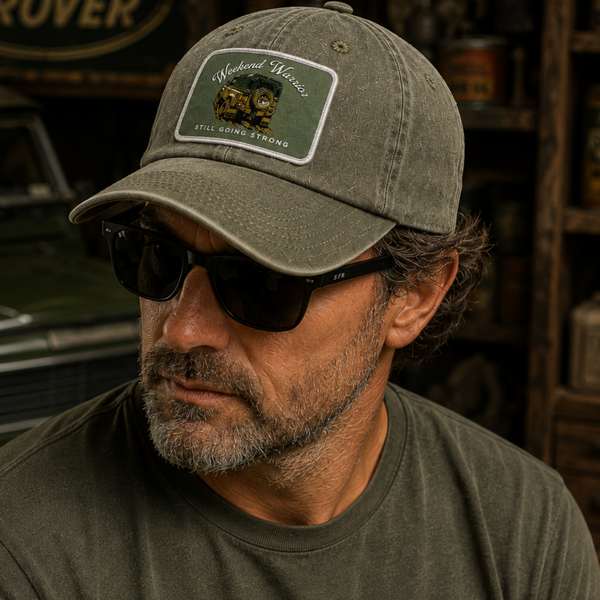 A man wearing a distressed olive green baseball cap with a 'Rancheria Ranchero' patch on the front, styled with dark sunglasses and an olive green t-shirt in a rustic, vintage-themed setting.